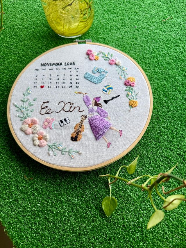 Hand embroidery: a hoop with a calendar, the name "Ee Xin", a girl playing violin and small flowers