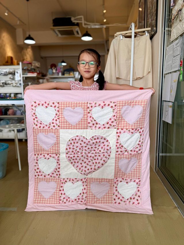 A young girl holding up a pink patchwork quilt with heart blocks