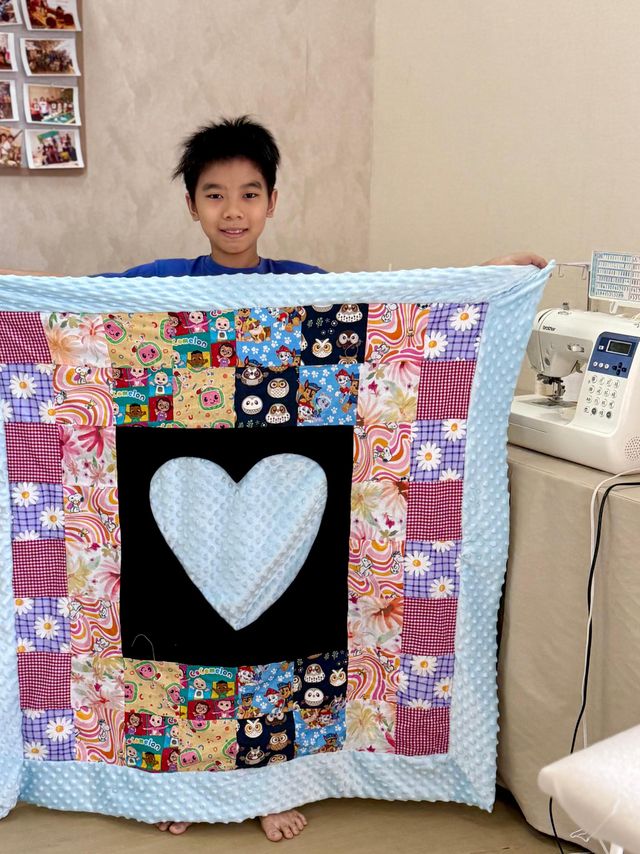 A boy holding up a colourful Bai Jia Bei patchwork quilt with a heart in the centre